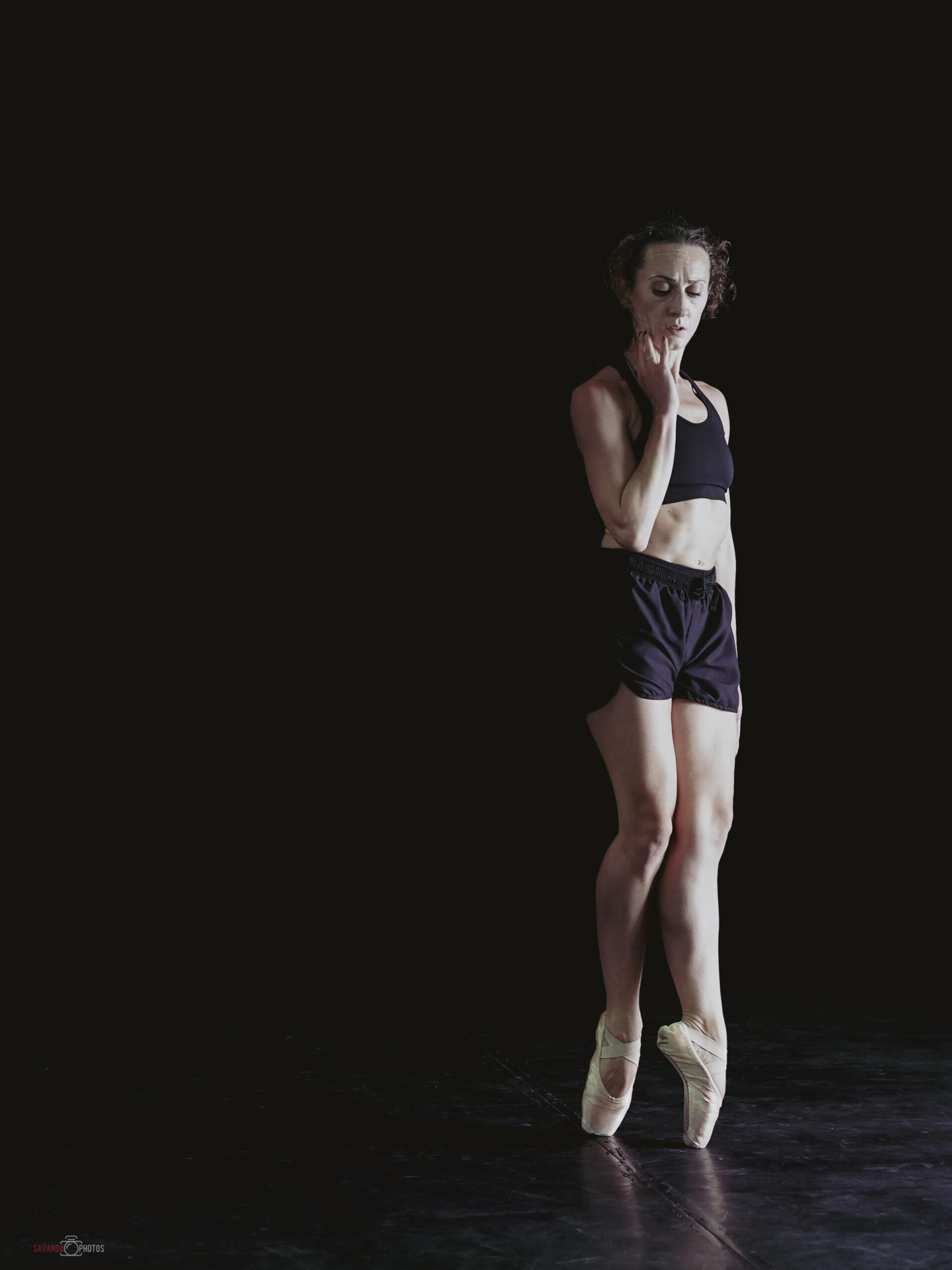 Chloé s'exécutant sur scène, vêtue d'un haut noir et de shorts, avec des pointes, se tenant dans une position contemplative sur fond sombre.